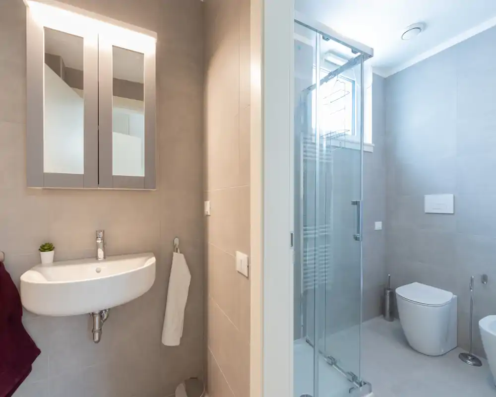 Modern, well-lit bathroom with a wall-mounted sink, mirrored cabinet, glass shower enclosure and a wall-hung toilet; neutral gray tiles create a clean, contemporary look.