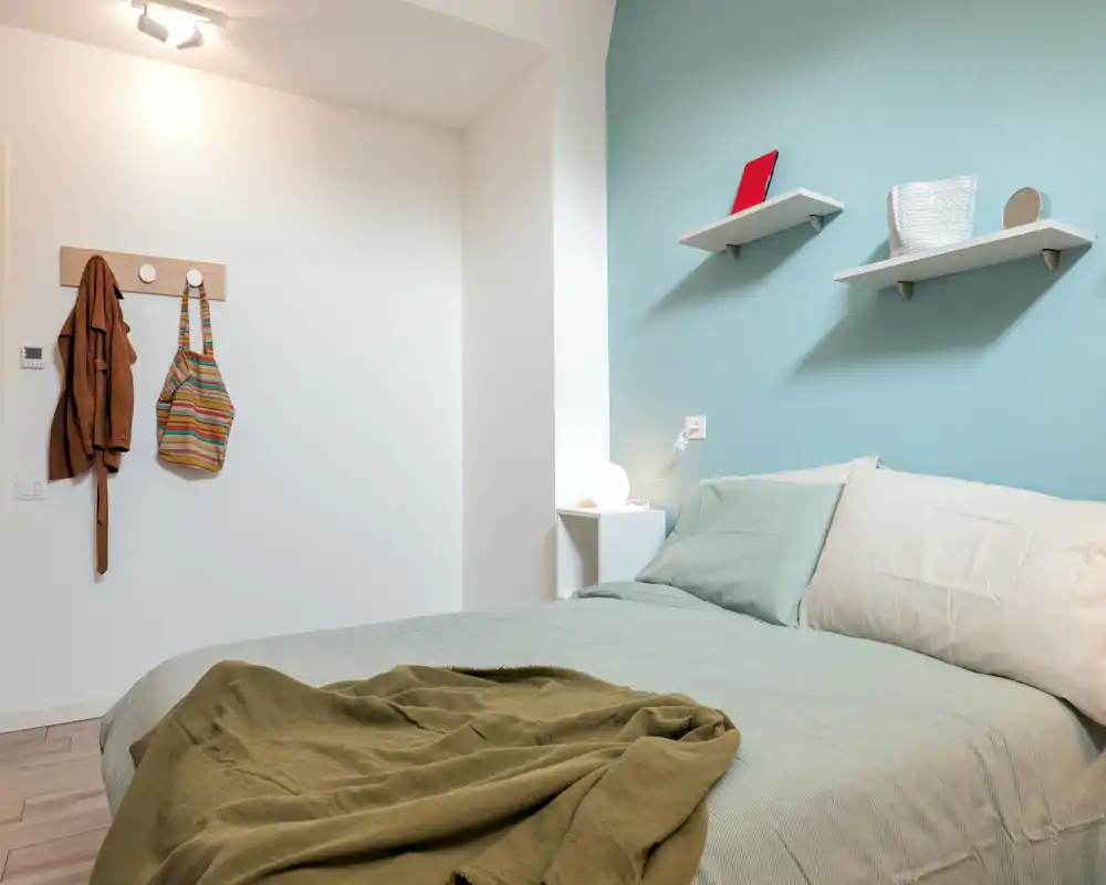 Bright, cozy bedroom with a neatly made double bed, soft green linens, bedside table with lamp and decorative shelves on a pastel blue wall.