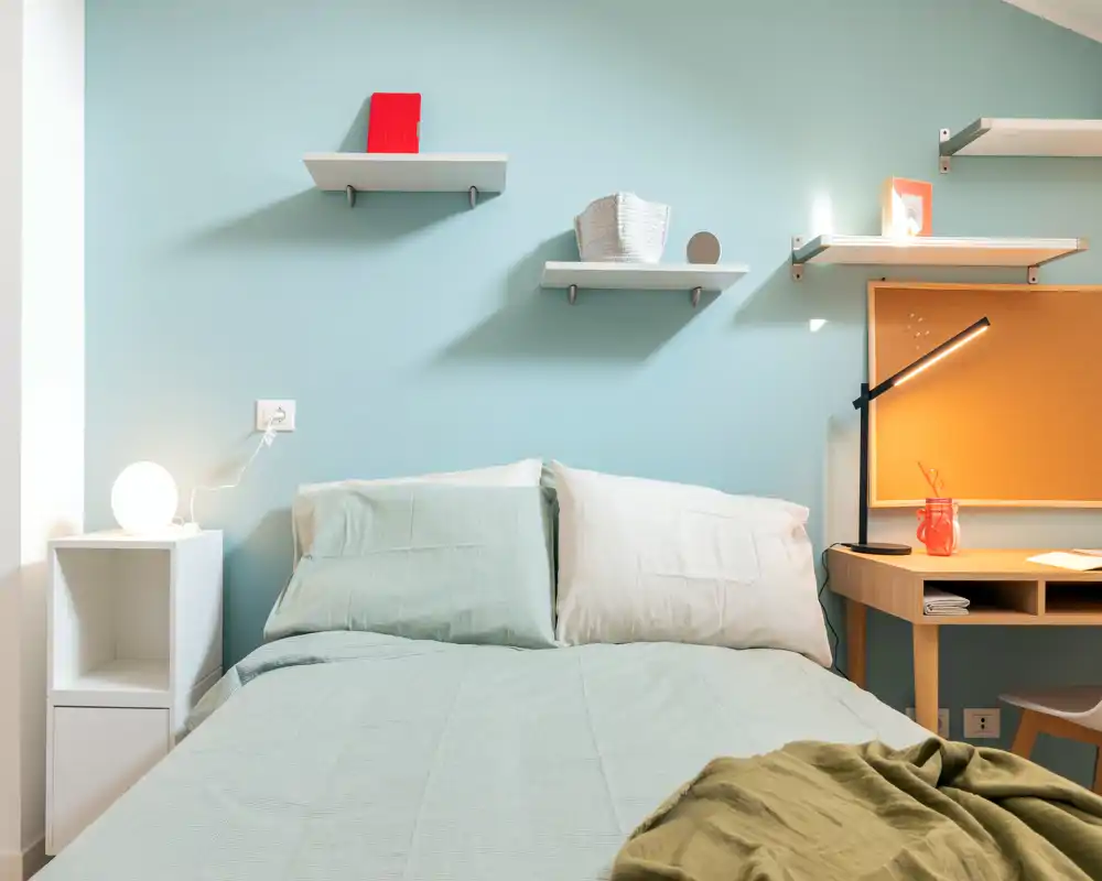 Bright, well-styled bedroom with a full bed centered in frame, soft pastel walls, bedside table and workspace with a desk lamp — ideal for single or couple stays.