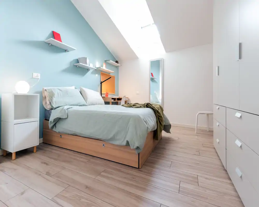 Bright, modern bedroom with a wooden platform bed, soft pastel bedding, and natural skylight. Clean, minimalist furnishings include a bedside table, wall shelves and a full-length mirror.