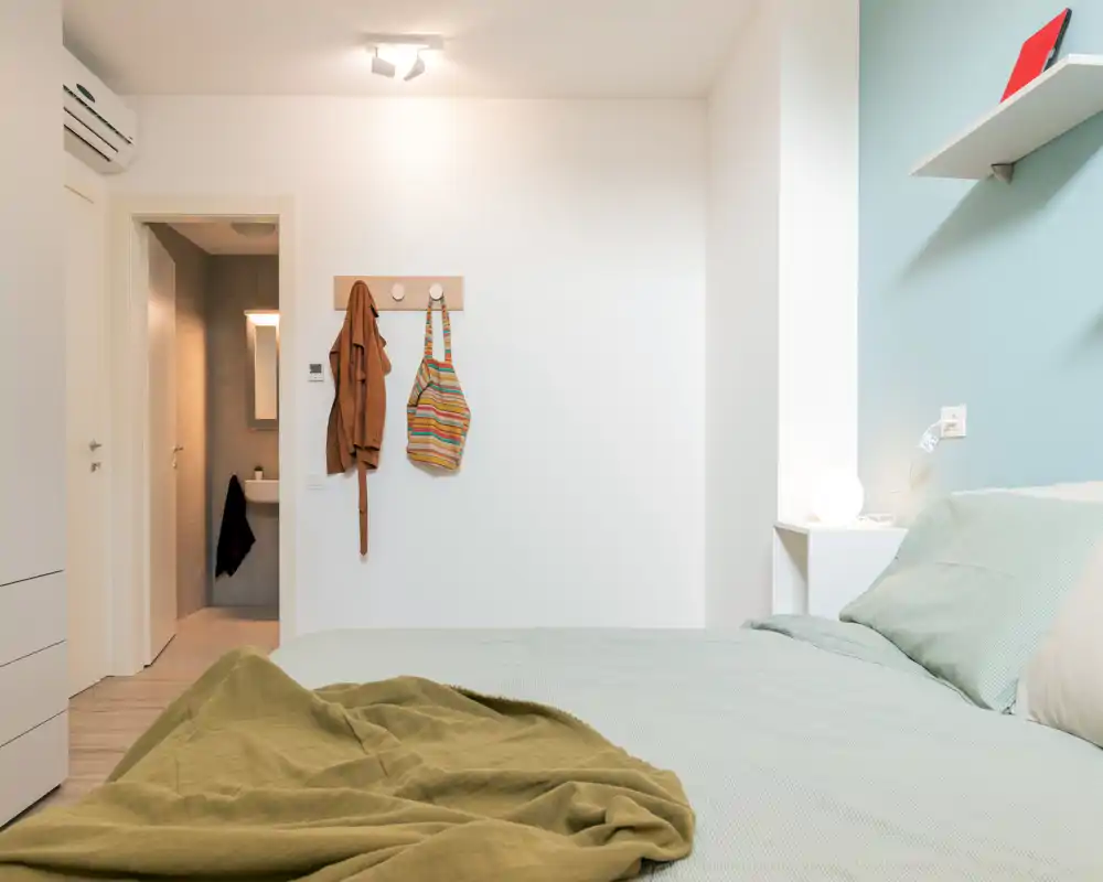 Light, modern bedroom with a neatly made double bed, soft green linens, bedside lamp and a teal accent wall. The composition highlights the sleeping area and a doorway to an en-suite bathroom.