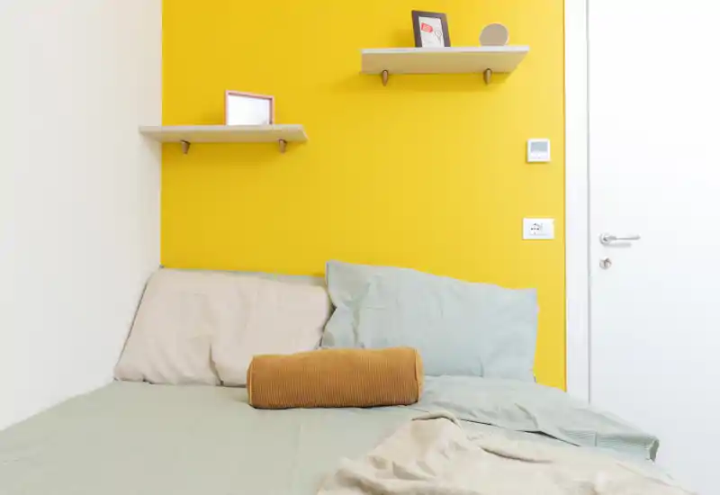 A compact, bright bedroom with a neatly made bed against a vibrant yellow accent wall, soft pillows and cozy textiles creating a welcoming nook.
