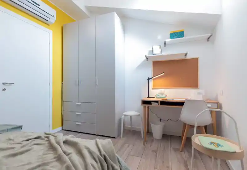 Modern, compact bedroom corner with a visible bed foreground, a tall wardrobe and a tidy wooden desk with task lamp — bright, clean and well organized for study and rest.