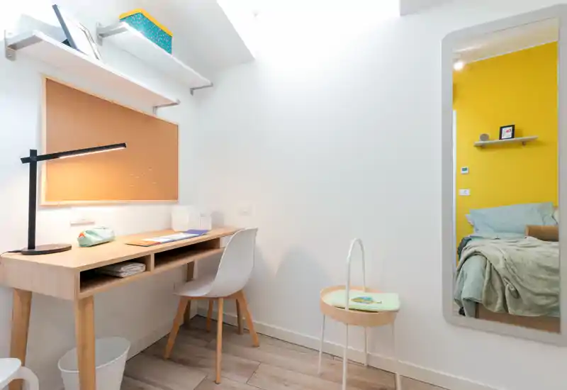 Compact bedroom corner with a study desk. Bright, modern space showing a wooden desk and chair in the foreground and a bed with soft textiles visible through the doorway; fresh white walls and a bold yellow accent add warmth.