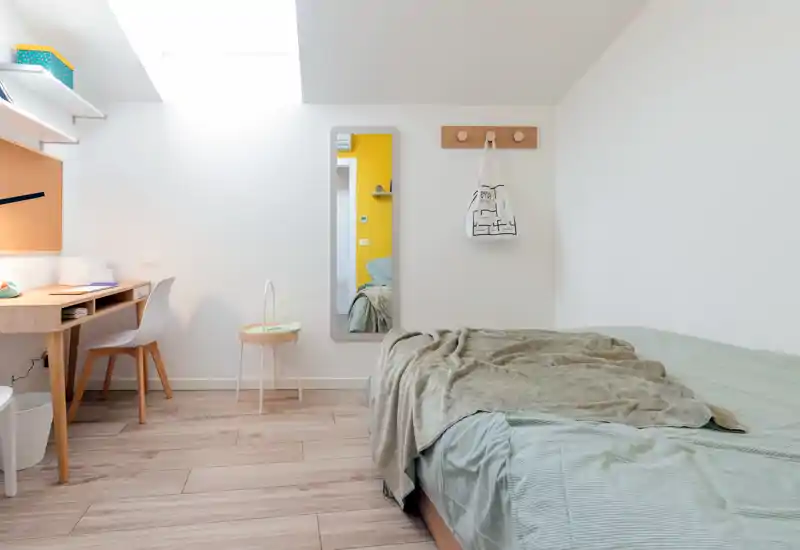 Bright, minimalist bedroom with a large bed dressed in soft green linens, a cozy throw, a wooden desk by the window and a full-length mirror—ideal for a calm, well-lit rental listing.