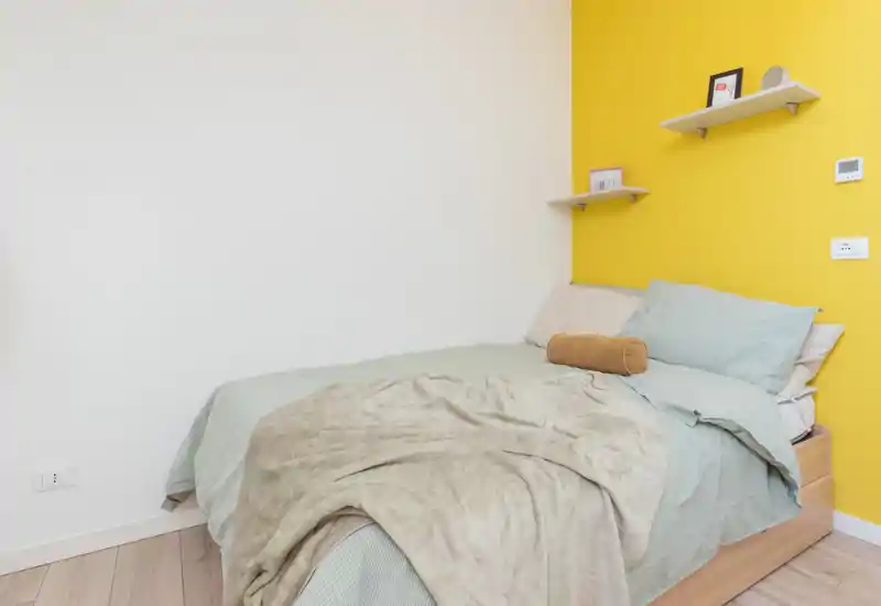 Bright, modern bedroom with a comfortable double bed against a yellow accent wall, soft linens and simple floating shelves for discreet storage.