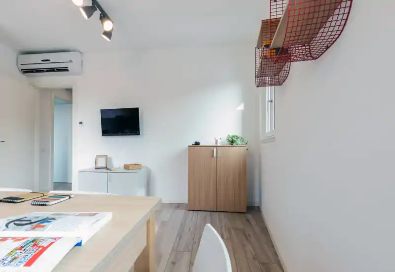 Bright, minimal living area with a dining/work table in the foreground, wall-mounted TV, compact storage units and a small window letting natural light in.