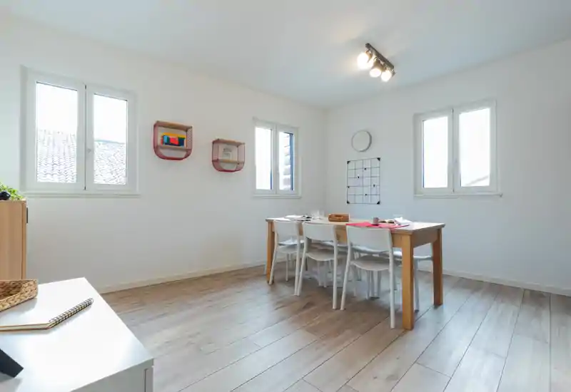 Bright, minimalist dining room with a wooden table and four white chairs, natural light from three windows and subtle wall decor.
