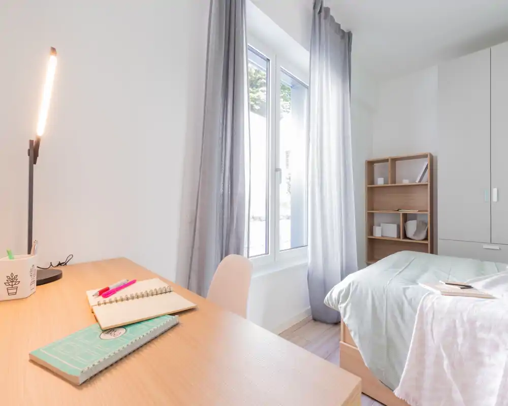 Camera da letto luminosa e moderna con letto singolo e biancheria morbida, scrittoio vicino alla finestra e una piccola libreria — perfetta per studenti o soggiorni brevi.