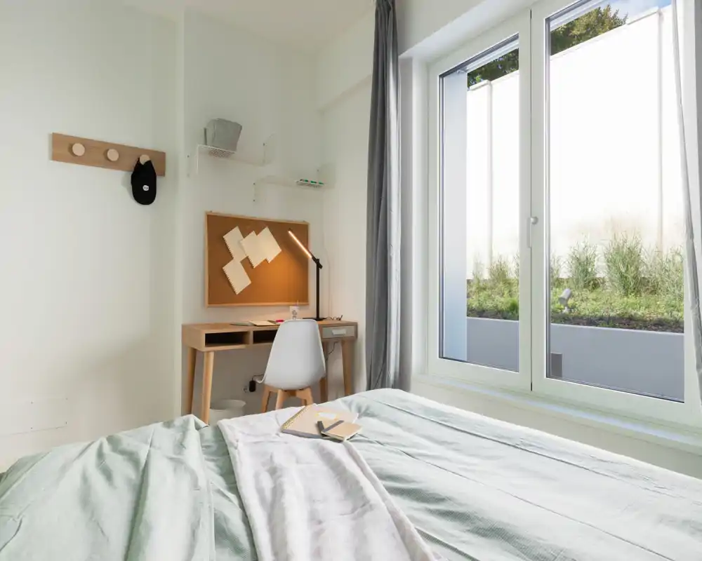 Camera da letto luminosa e minimal con letto ben rifatto in primo piano, una piccola scrivania in legno e una sedia vicino a una grande finestra che offre molta luce naturale.