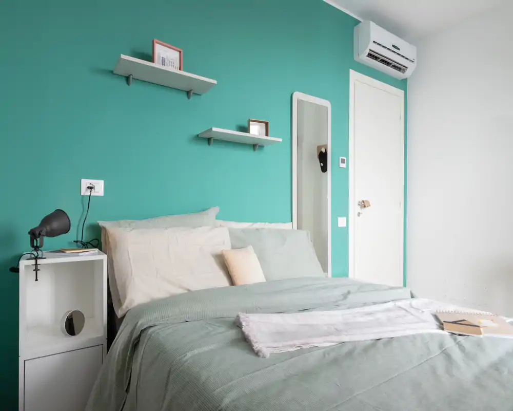 Camera da letto moderna e luminosa con letto matrimoniale ben rifatto davanti a una parete accentata turchese, comodino e mensole per un'atmosfera tranquilla e di stile.