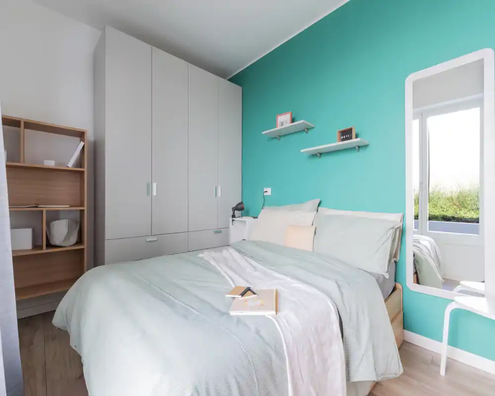 Camera da letto moderna e luminosa con letto matrimoniale, biancheria morbida e parete accentata turchese; armadio a muro e specchio che creano un'atmosfera accogliente.