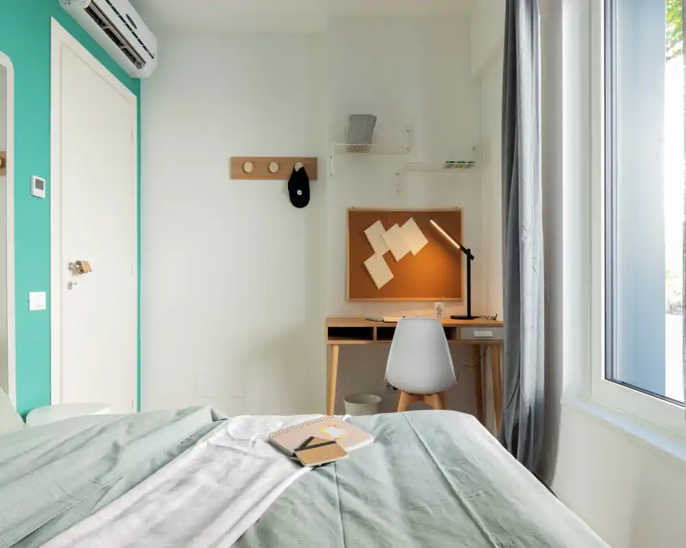 Camera da letto moderna e luminosa con letto ordinato, piccolo scrittoio e ampia finestra che illumina la stanza — ambiente minimal e ben allestito per annunci.