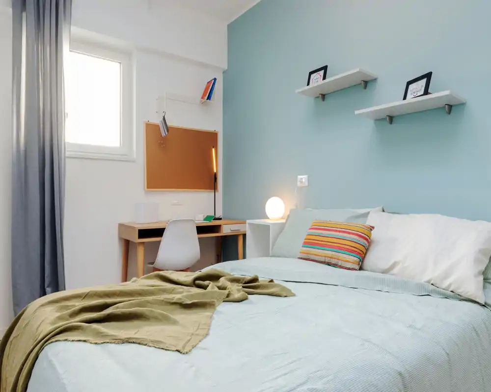 Dormitorio moderno y luminoso con cama grande, lámpara de mesita y pequeño escritorio — decorado con cojín a rayas y manta, perfecto para un anuncio.