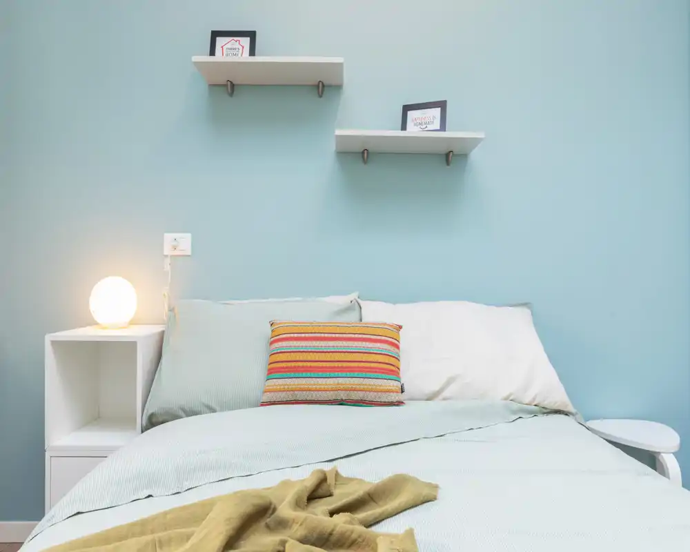 Dormitorio bien decorado con cama doble cómoda, ropa de cama en tonos pastel, cojines decorativos y una lámpara de mesita cálida frente a una pared azul claro.