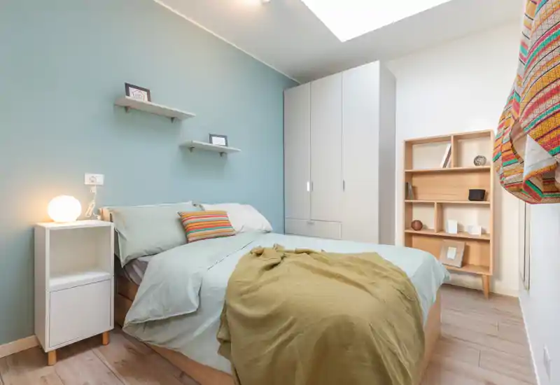 Camera da letto luminosa e accogliente con comodo letto matrimoniale, biancheria morbida, comodino con lampada, armadio e scaffalatura — perfetta per un soggiorno rilassante.