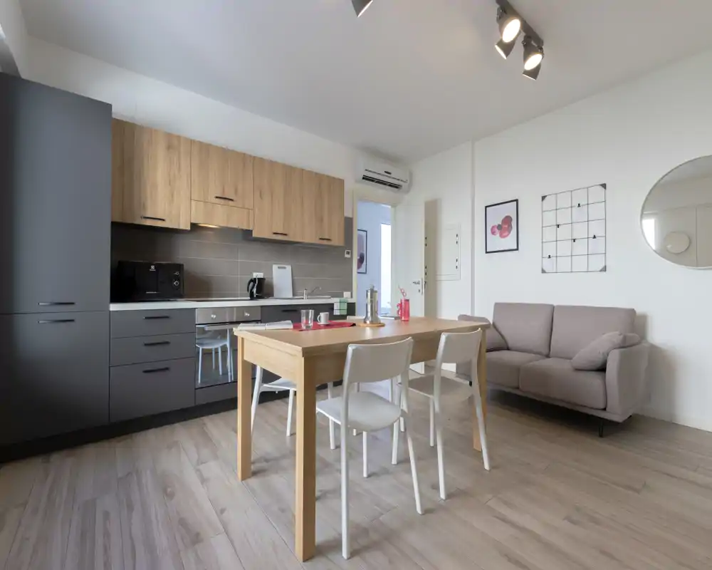 Espacio de estar moderno y luminoso tipo open-plan con cocina integrada y mesa de madera; un sofá compacto y decoración de pared que hacen el ambiente acogedor.