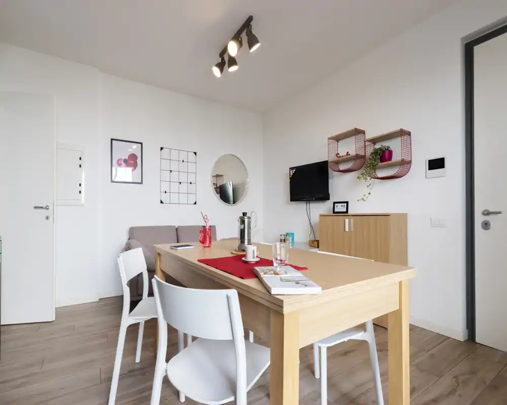 Estancia luminosa y moderna con mesa de madera, sillas blancas, pequeño sofá y televisor en la pared — espacio cuidado e ideal para alquileres de corta estancia.