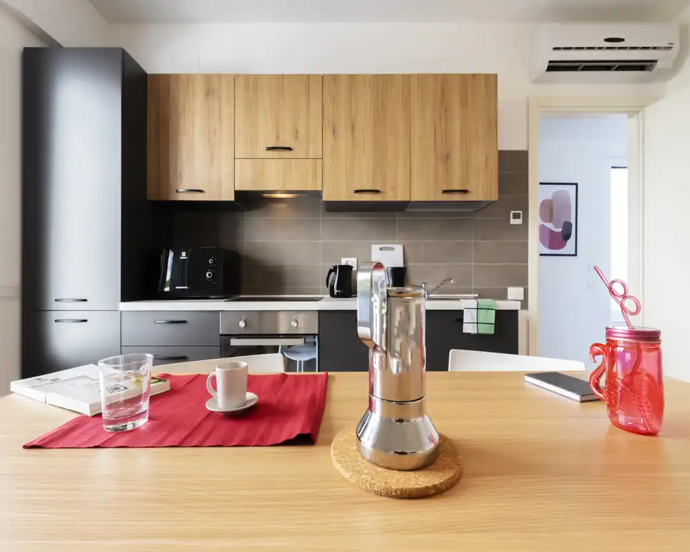 Cocina moderna y luminosa con armarios de madera, horno empotrado y encimera con cafetera moka y disposición informal en la mesa—perfecta para un anuncio de alquiler.
