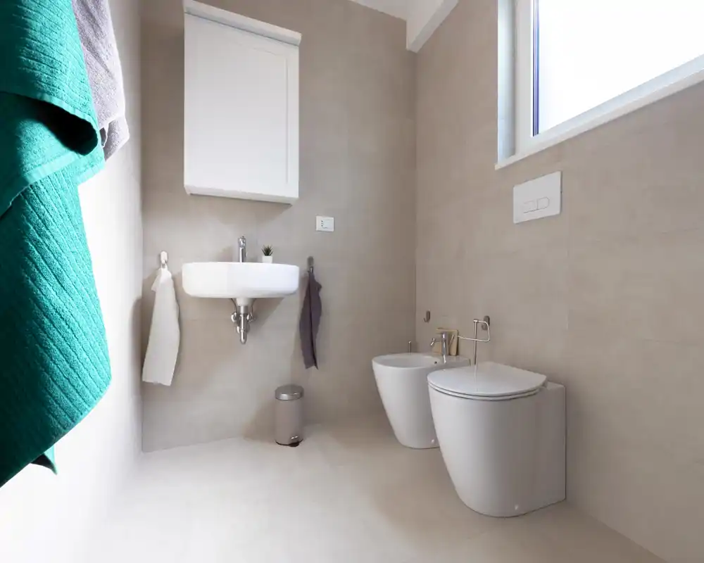 Baño moderno y luminoso con lavabo mural, bidé e inodoro; azulejos neutros y ventana que aportan luz natural para un aspecto limpio y minimalista.