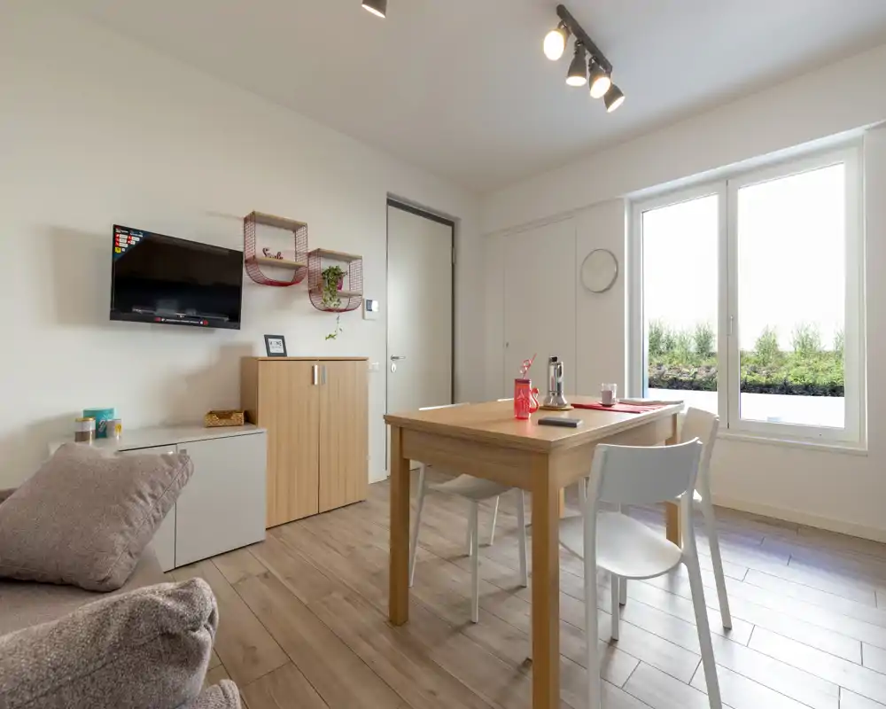 Zona de estar moderna y luminosa con mesa de comedor de madera, sillas blancas y un sofá cómodo. Una gran ventana inunda la habitación de luz natural y la decoración sencilla transmite calidez.