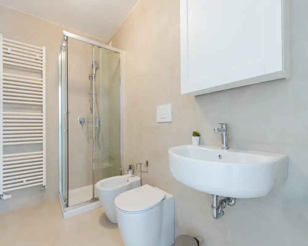 Baño moderno y compacto con mampara de ducha de cristal, lavabo empotrado en pared, inodoro y bidé sobre azulejos claros.