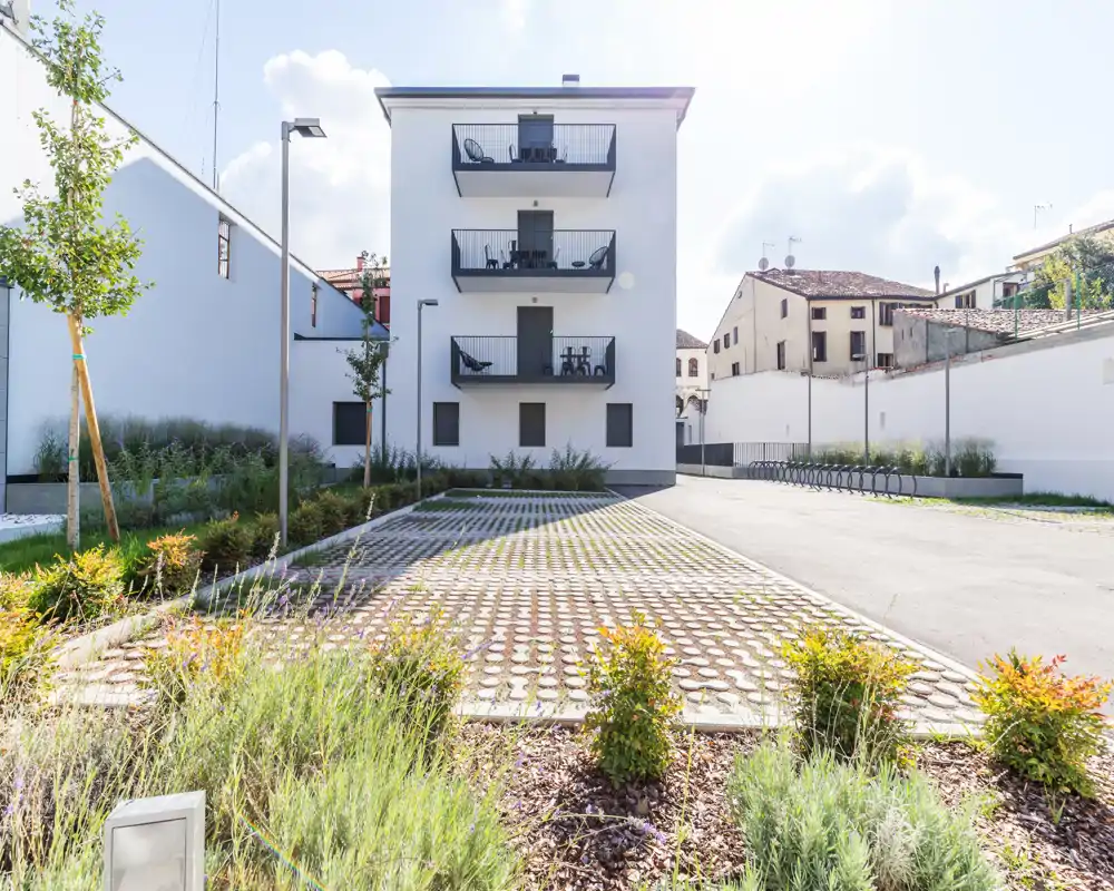 Patio/área de aparcamiento soleada frente a un edificio residencial moderno con balcones y parterres ajardinados, ideal para mostrar el espacio exterior y la fachada.