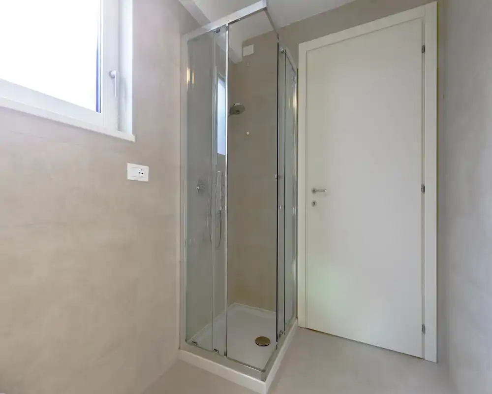 Baño moderno y compacto con ducha cerrada de cristal, azulejos en tonos neutros y ventana translúcida que aporta luz natural. Líneas limpias y diseño minimalista para un espacio funcional y atractivo.