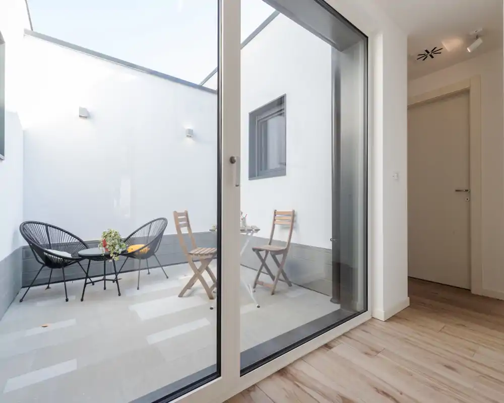 Pequeño patio privado visible a través de una gran puerta de vidrio, amueblado con dos sillas tejidas negras y una pequeña mesa además de dos sillas plegables de madera — zona exterior luminosa y minimalista ideal para el café matutino.