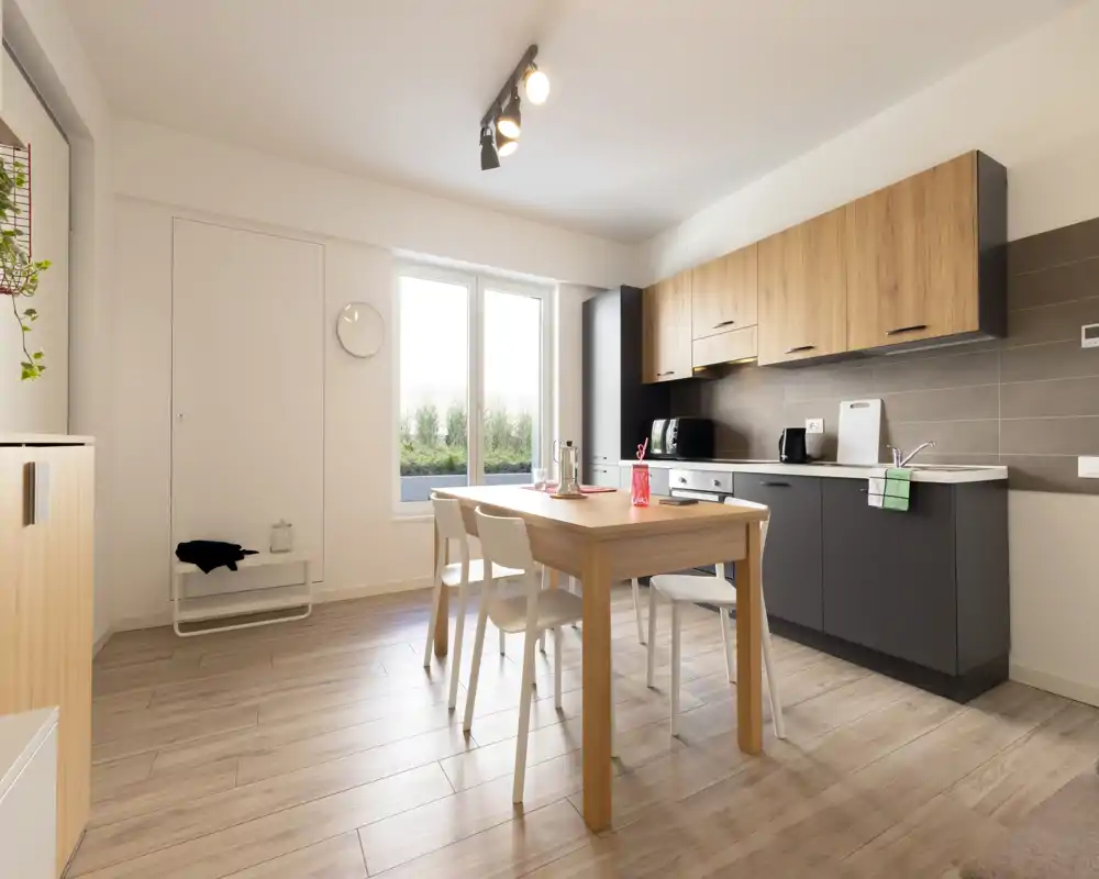 Cocina moderna y luminosa con muebles de madera y gris, mesa de comedor central de madera y sillas, con luz natural de una ventana grande.