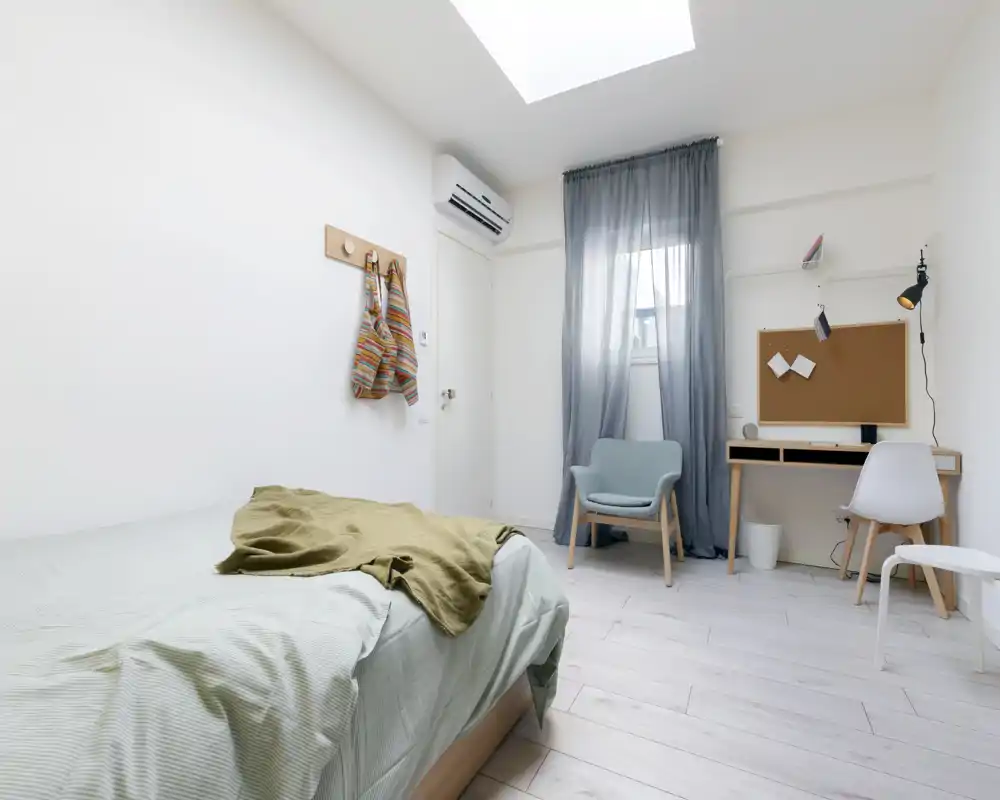 Chambre lumineuse et minimaliste avec un lit simple, un plaid confortable et un petit espace bureau près de la fenêtre. La pièce dispose d'une fenêtre de toit, de tons neutres et d'un fauteuil cosy, idéale pour une annonce locative.