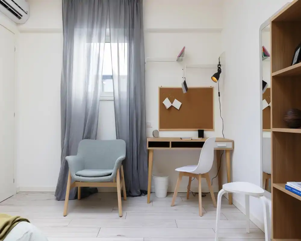 Coin de chambre lumineux et minimaliste avec un petit espace bureau : fauteuil doux, bureau avec panneau en liège et chaise, rideaux clairs et sol en bois pâle pour une ambiance apaisante.