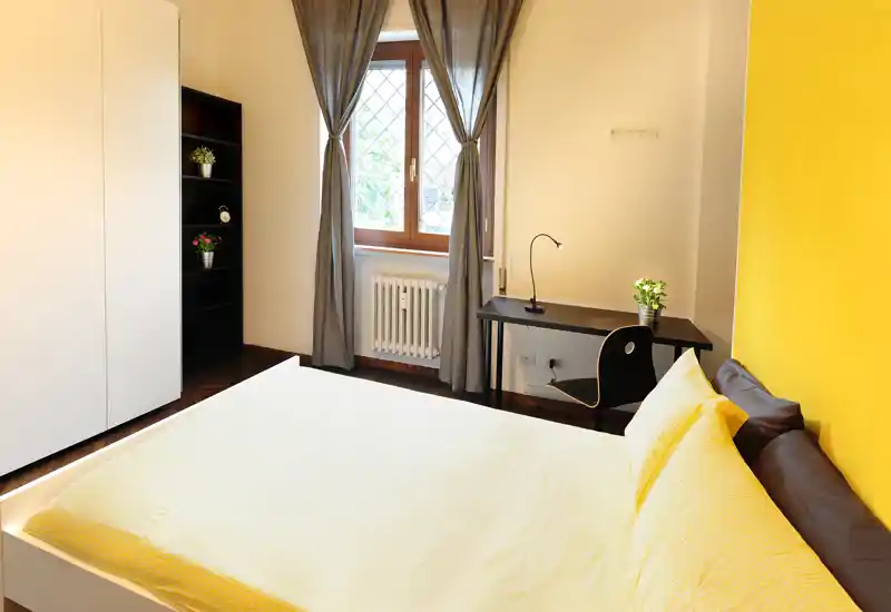 Camera da letto luminosa e moderna con un grande letto con biancheria gialla, una scrivania vicino alla finestra e un armadio. La luce naturale e la parete gialla donano all'ambiente un aspetto fresco e accogliente.