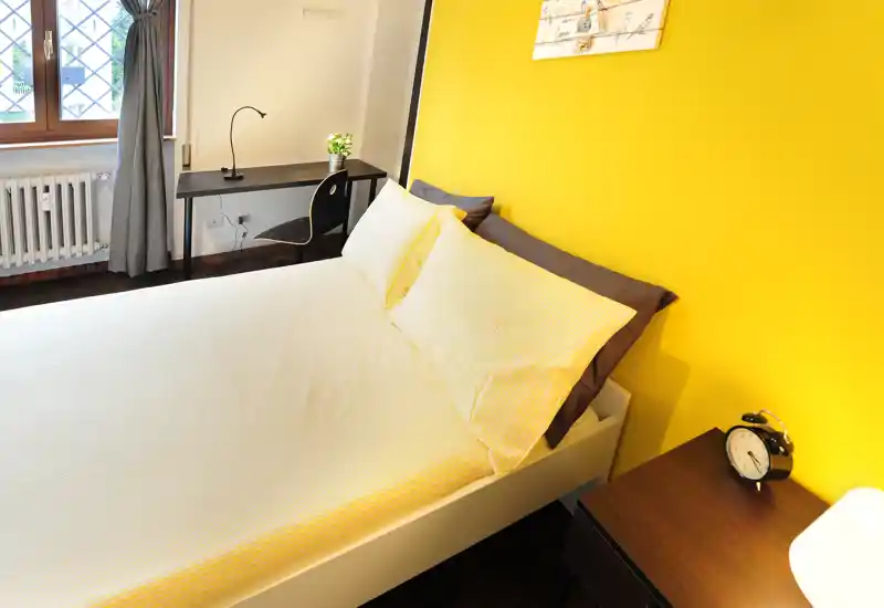 Camera da letto moderna e luminosa con un grande letto con lenzuola giallo pallido contro una parete accentuata di giallo vivace. Un comodino con sveglia e una piccola scrivania vicino alla finestra offrono una disposizione pratica e accogliente, ideale per affitti brevi.