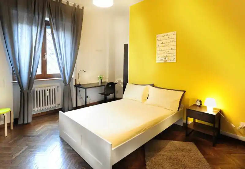 Camera da letto luminosa e ben arredata con letto matrimoniale bianco contro una parete gialla vivace, comodino e scrivania vicino alla finestra — ideale per annunci in affitto che valorizzano un ambiente accogliente e moderno.
