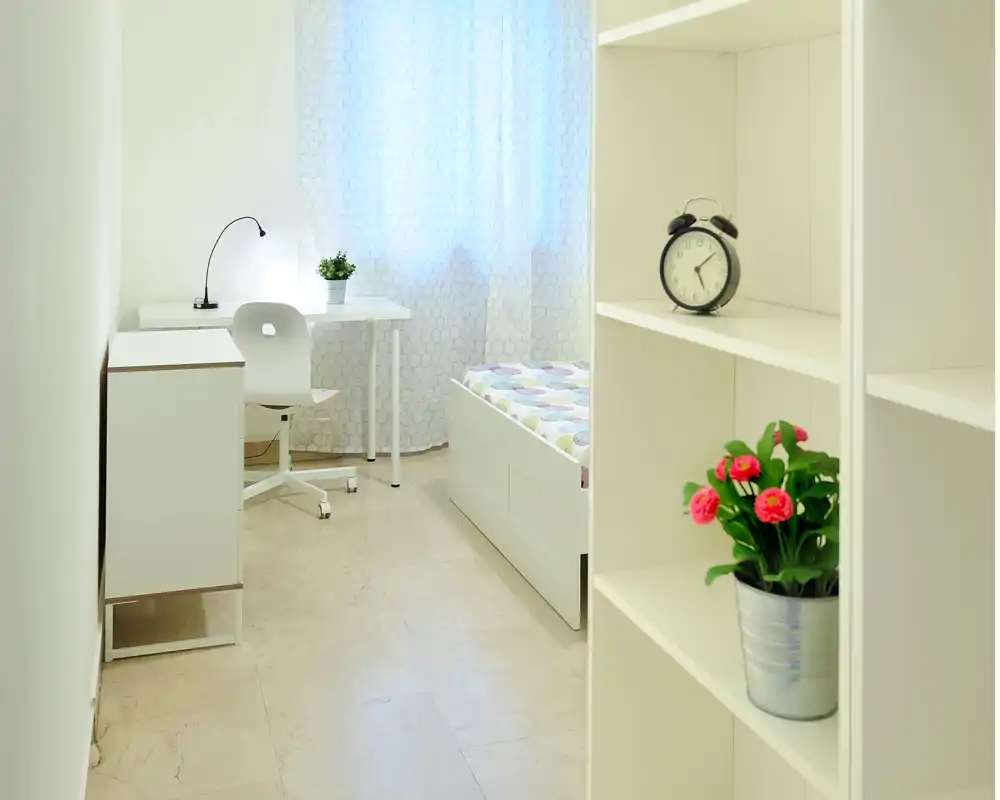 Camera da letto luminosa e minimal con letto singolo, piccolo scrittoio e scaffalatura — stile pulito e semplice ideale per studenti o ospiti.