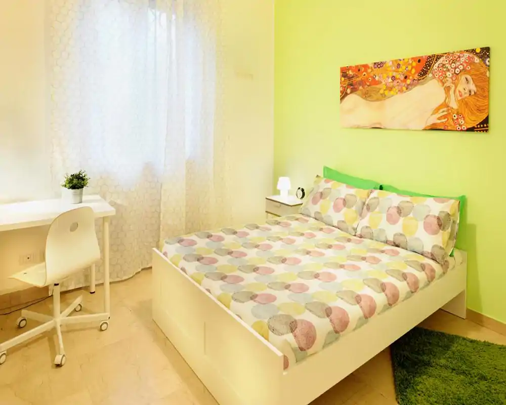 Camera da letto luminosa e moderna con letto matrimoniale, biancheria a fantasia, comodino con lampada e una piccola scrivania vicino alla finestra — pulita, accogliente e ben illuminata.