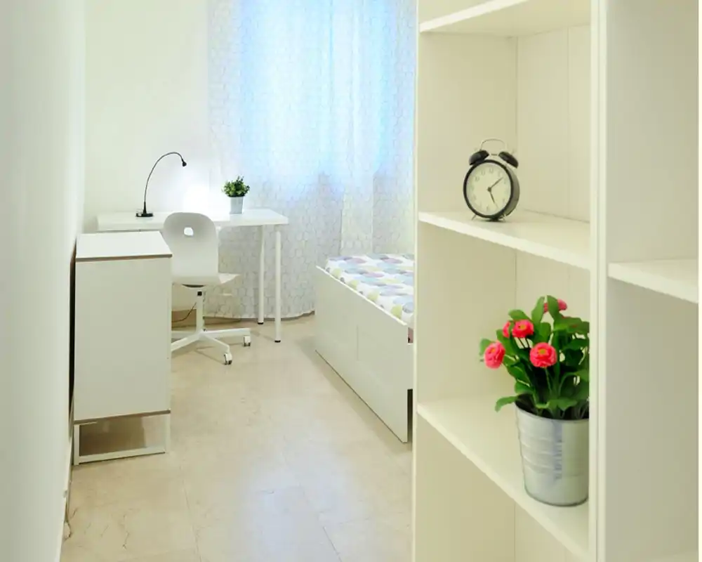 Camera da letto luminosa e minimal con letto singolo, scrivania e mensole; decorazioni bianche, luce naturale e piccole piante donano un’atmosfera fresca e ariosa.