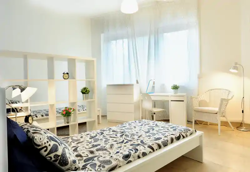 Dormitorio luminoso y bien decorado con cama individual en primer plano, estantería divisoria blanca, escritorio junto a la ventana y sillón acogedor — perfecto para un anuncio que destaque la luz y el confort.