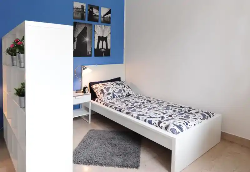 Dormitorio individual moderno y luminoso con cama blanca bien hecha, ropa de cama estampada, mesita con lámpara y estantería que divide parcialmente el espacio con plantas decorativas.