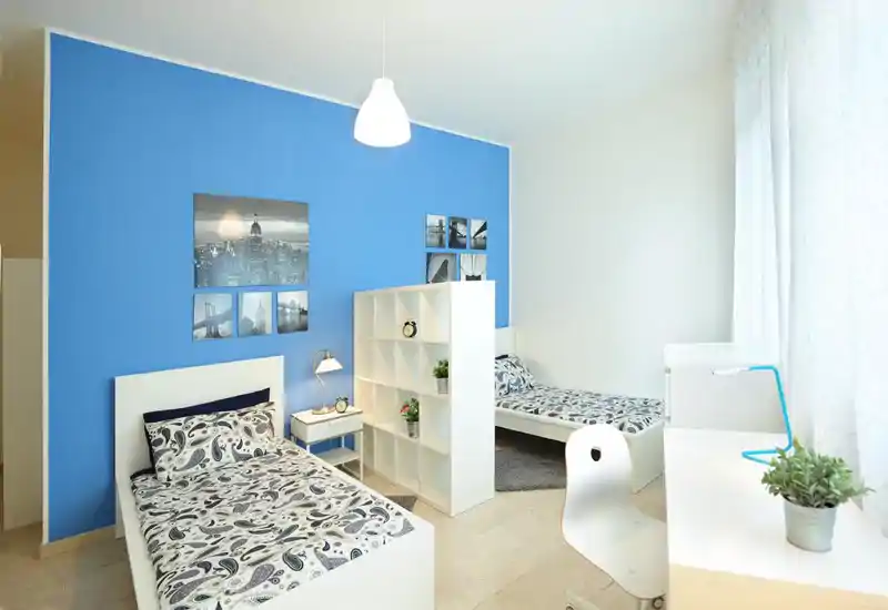 Camera da letto doppia moderna e luminosa con due letti singoli separati da un divisorio a cubi, biancheria con motivo, scrivania vicino alla finestra e parete azzurra come accento vivace.