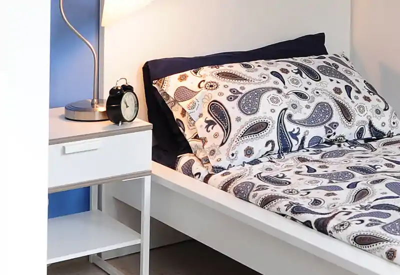Rincón de dormitorio individual luminoso y bien dispuesto con ropa de cama estampada, mesita, lámpara y despertador — ideal para un anuncio de alquiler.