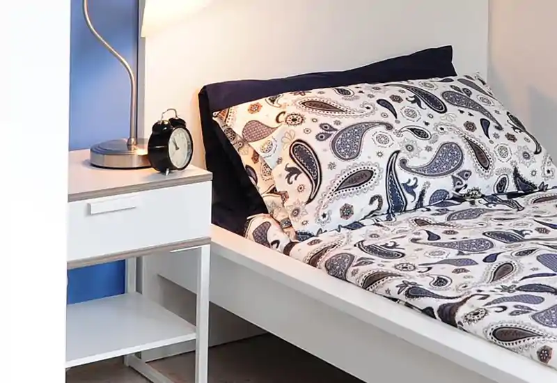 Angolo camera luminoso e ordinato con letto bianco e biancheria stampata, comodino, lampada e sveglia — un ambiente notte accogliente e ben illuminato, perfetto per annunci.