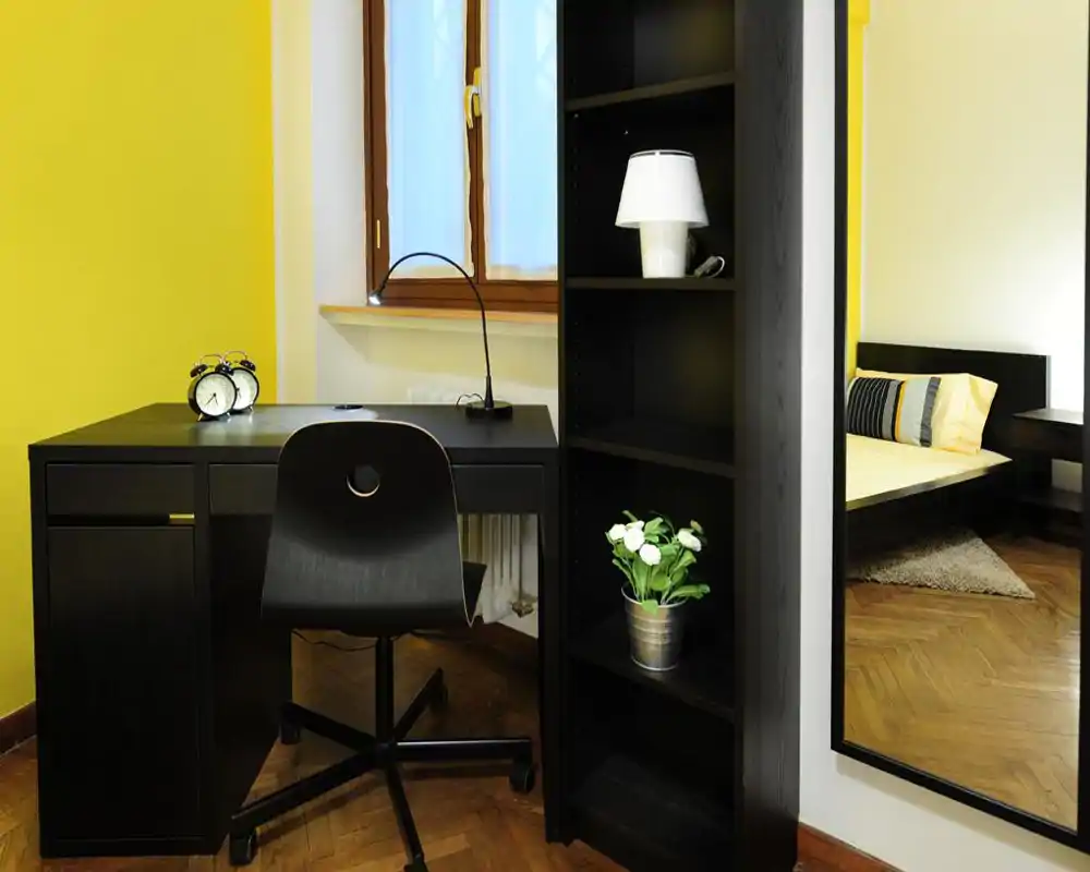 Rincón de dormitorio luminoso y moderno con cama visible, textiles acogedores y un escritorio. La imagen destaca las zonas de descanso y trabajo con paredes amarillas y mobiliario contemporáneo.