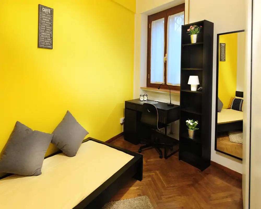 Dormitorio individual luminoso y acogedor con pared amarilla, cama con cojines, escritorio junto a la ventana y una estantería alta con espejo — ideal para estudiante o viajero solo.