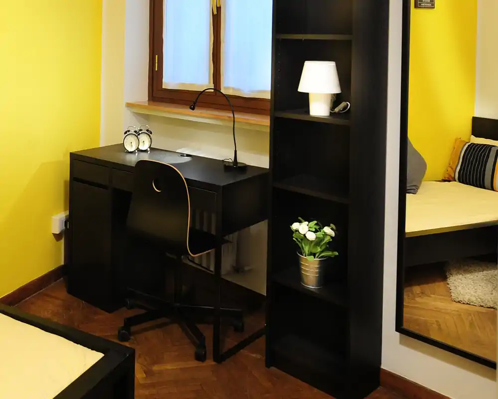 Rincón de dormitorio bien decorado con cama visible, escritorio junto a la ventana, estantería y paredes amarillas cálidas que generan una atmósfera acogedora.
