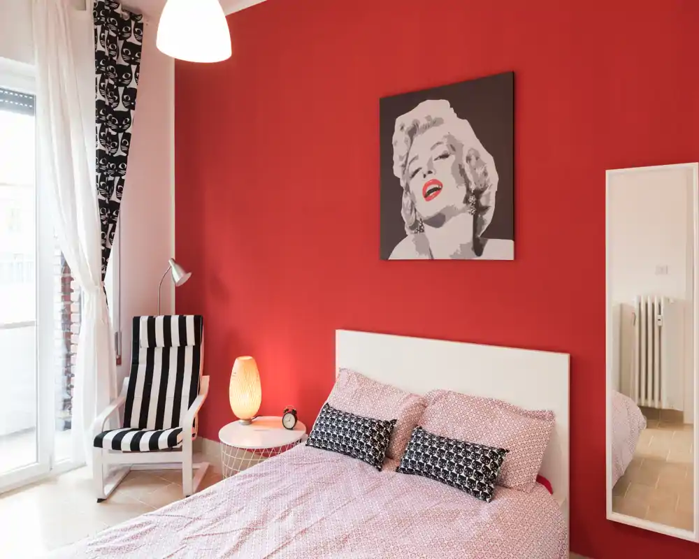 Bright, stylish bedroom with a full bed against a bold red feature wall, decorative cushions, a reading chair by the window and modern artwork above the headboard.