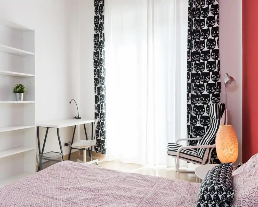 Bright, stylish bedroom with patterned curtains and a comfortable double bed in the foreground, a small desk by the window and a cozy reading chair with a lamp.