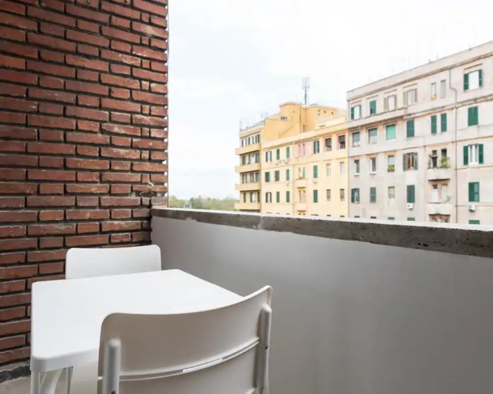 Small urban balcony with a white plastic table and chairs, a brick accent wall and views over neighboring apartment buildings — bright, simple outdoor seating ideal for morning coffee.