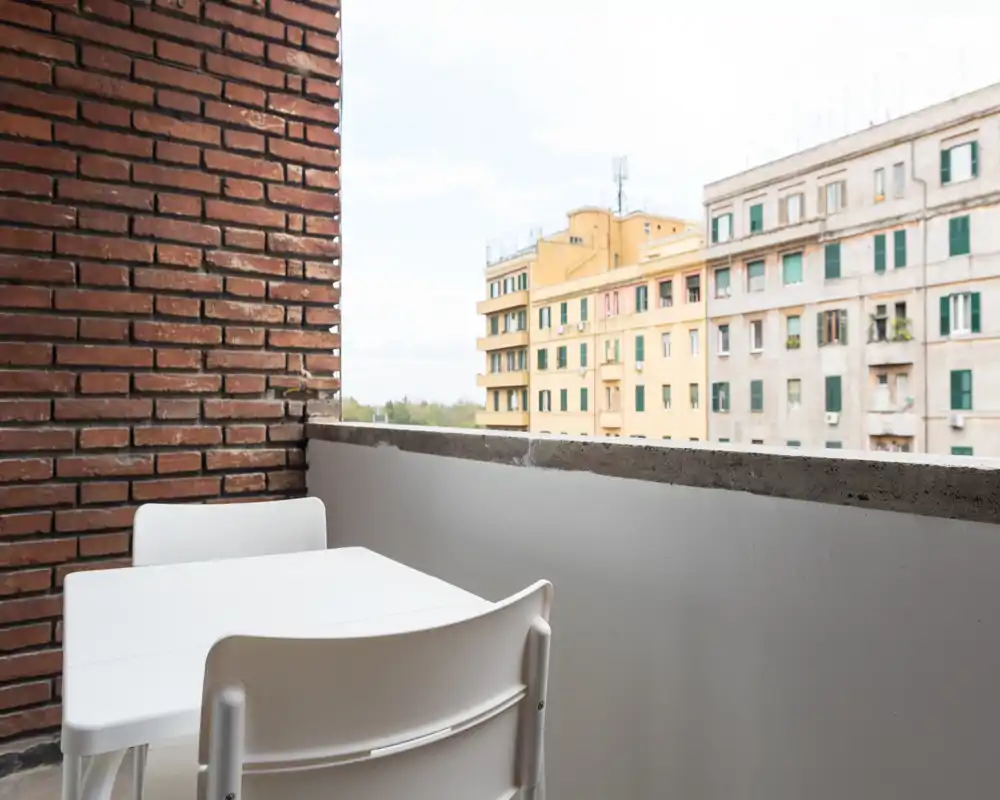 Small urban balcony with a white table and chairs, exposed brick wall at left and views over neighboring apartment buildings — bright, functional outdoor seating.